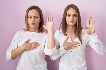 Middle age mother and young daughter standing over pink background swearing with hand on chest and...
