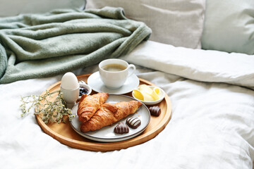 Mothers day breakfast served with love on bed. Coffee, croissant, egg and chocolate hearts on a wooden tray. Natural bed cloth. Also for birthday, valentines or fathers day, copy space - obrazy, fototapety, plakaty