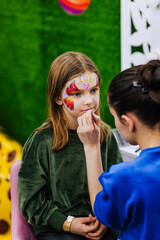 A professional make-up artist, artist paints with a brush on her face with face paints, drawing, children's makeup for a little girl model, a child. Photography, art, concept, lifestyle.