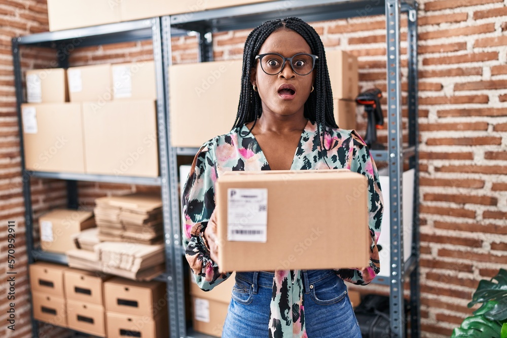 Sticker african woman with braids working at small business ecommerce holding package in shock face, looking