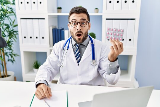 Handsome hispanic man wearing doctor uniform holding prescription pills scared and amazed with open mouth for surprise, disbelief face