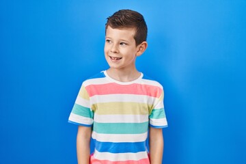 Young caucasian kid standing over blue background looking away to side with smile on face, natural expression. laughing confident.