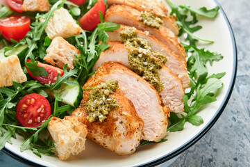 Fresh salad with grilled chicken fillet, arugula, tomatoes, cucumber and pesto sauce, sesame seeds with olive oil in white bowl on light slate background. Healthy lunch menu. Diet food. Top view.