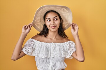 Young brazilian woman wearing summer hat over yellow background smiling looking to the side and staring away thinking.