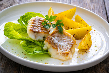 Fish dish - fried cod with baked potatoes and fresh vegetables on wooden table
