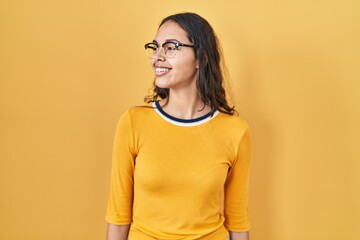 Obraz premium Young brazilian woman wearing glasses over yellow background looking away to side with smile on face, natural expression. laughing confident.