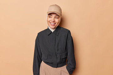 Horizontal shot of happy Islamic woman wears hijab and black jacket smiles broadly looks aside smiles joyfully being in good mood isolated over brown background. Stylish Muslim female model.