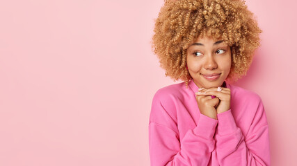 Horizontal shot of pretty thoughtful curly haired keeps hands under chin smiles gently focused away dressed in casual sweatshirt isolated over pink background copy space for your promotional content