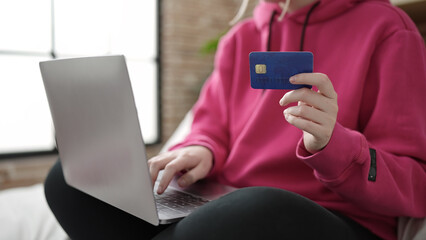 Young blonde woman using laptop and credit card sitting on bed at bedroom