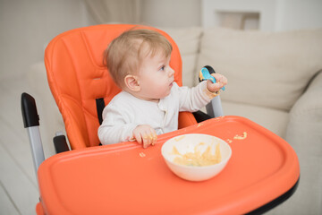 a cute boy sitting on a chair holds a spoon in his hand and eats baby vegetable puree himself. Healthy food for a child. Child eats independently
