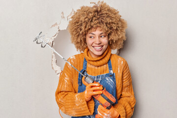 Cheerful female plasterer holds mixer for mixing plaster on walls wears orange jumper and...