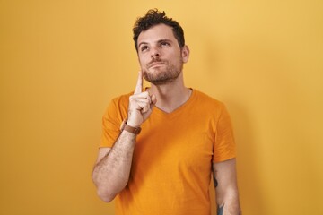 Young hispanic man standing over yellow background thinking concentrated about doubt with finger on chin and looking up wondering