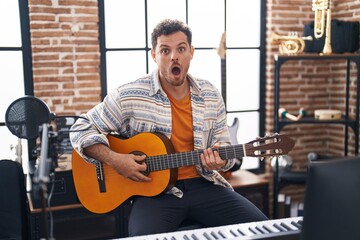 Young hispanic man playing classic guitar at music studio scared and amazed with open mouth for surprise, disbelief face