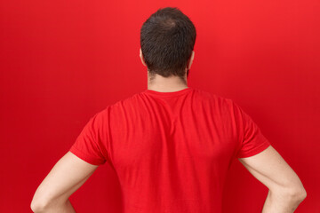 Young hispanic man wearing casual red t shirt standing backwards looking away with arms on body