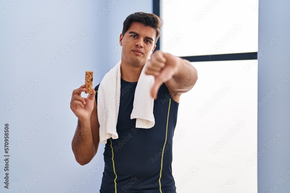 Poster Hispanic man eating protein bar as healthy energy snack with angry face, negative sign showing dislike with thumbs down, rejection concept