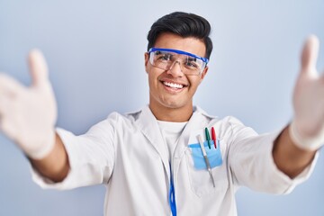Hispanic man working as scientist looking at the camera smiling with open arms for hug. cheerful expression embracing happiness.