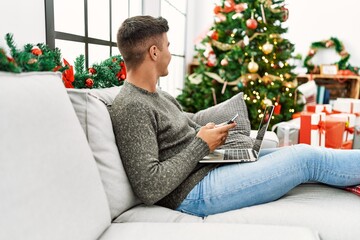 Young hispanic man using laptop and smartphone sitting on sofa by christmas tree at home