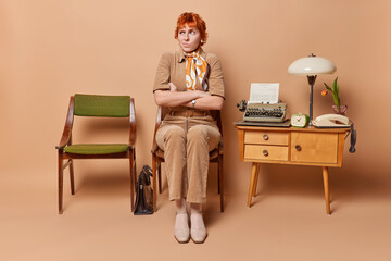 Pensive young redhead vintage woman poses in retro room with typewriter on table sits on old chair keeps arms folded dressed in elegant clothes isolated over brown background. Mid 20th century