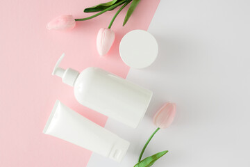 Obraz na płótnie Canvas Natural skin care products concept. Top view photo of white cosmetic bottles, cream jar and spring flowers on pastel pink and white background. Flat lay cosmetic mockup.