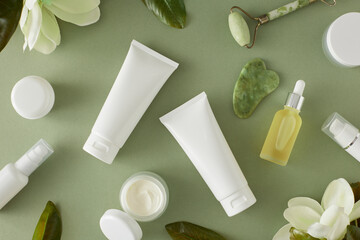 Organic cosmetic concept. Flat lay mockup of white cream tubes, dropper bottles, cream jars, face massage roller and tropical flowers on pastel green background.
