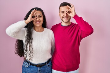 Young hispanic couple standing over pink background doing ok gesture with hand smiling, eye looking through fingers with happy face.