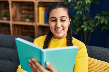 Young hispanic woman reading book sitting on sofa at home