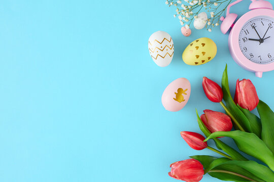Easter holiday concept. Flat lay photo of color eggs, tulips flowers and  pink alarm clock on pastel blue background with copyspace. Easter coming idea.
