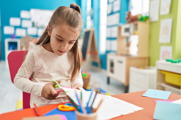 Adorable hispanic girl student sitting on table cutting paper at kindergarten