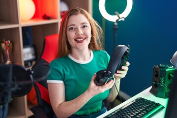 Young redhead woman streamer smiling confident holding headphones at gaming room