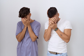Homosexual gay couple standing over white background with sad expression covering face with hands...