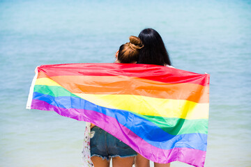 LGBTQI couple on the beach with rainbow flag beautiful female young lesbian couple in love holding hands, and a rainbow flag, a symbol of the LGBT community, equal rights, beauty and love