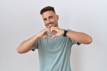 Hispanic man with beard standing over white background smiling in love doing heart symbol shape with hands. romantic concept.