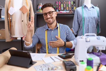 Hispanic man with beard dressmaker designer working at atelier smiling swearing with hand on chest...