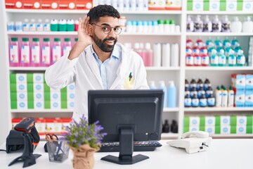 Hispanic man with beard working at pharmacy drugstore smiling with hand over ear listening an hearing to rumor or gossip. deafness concept.