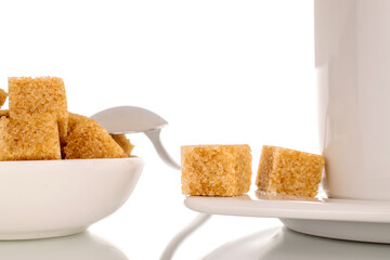 Several cubes of brown sugar with ceramic white cup with saucer and metal spoon, macro, isolated on white background.