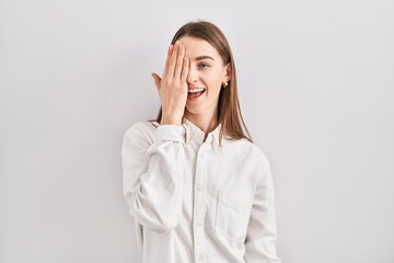 Young caucasian woman standing over isolated background covering one eye with hand, confident smile on face and surprise emotion.