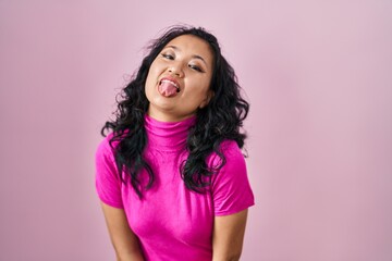 Young asian woman standing over pink background sticking tongue out happy with funny expression....