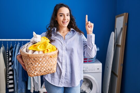 Young brunette woman holding laundry basket surprised with an idea or question pointing finger with happy face, number one