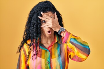 Middle age hispanic woman standing over yellow background peeking in shock covering face and eyes with hand, looking through fingers with embarrassed expression.