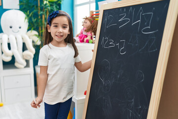 Adorable hispanic girl preschool student smiling confident writing numbers on blackboard at...