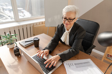 Confident stylish european middle aged senior woman using laptop at workplace. Stylish older mature 60s gray haired lady businesswoman sitting at office table. Boss leader teacher professional worker.