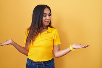 Young arab woman standing over yellow background smiling showing both hands open palms, presenting...