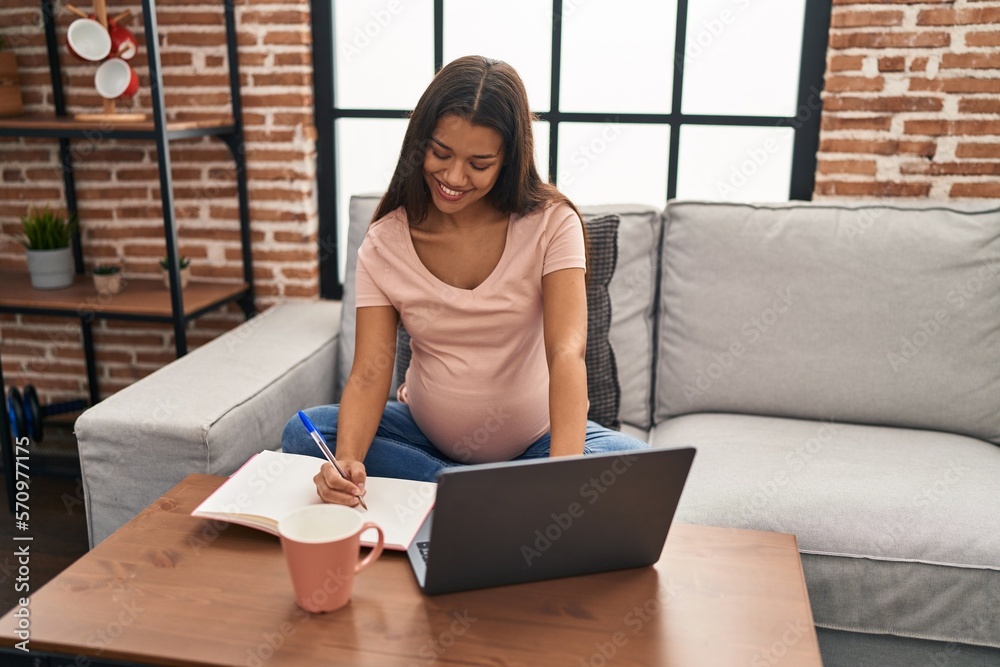 Sticker young latin woman pregnant using laptop writing on book at home