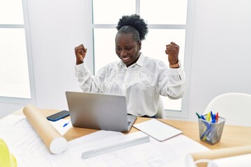 Young african american woman architect using laptop with fists raised up at architecture studio