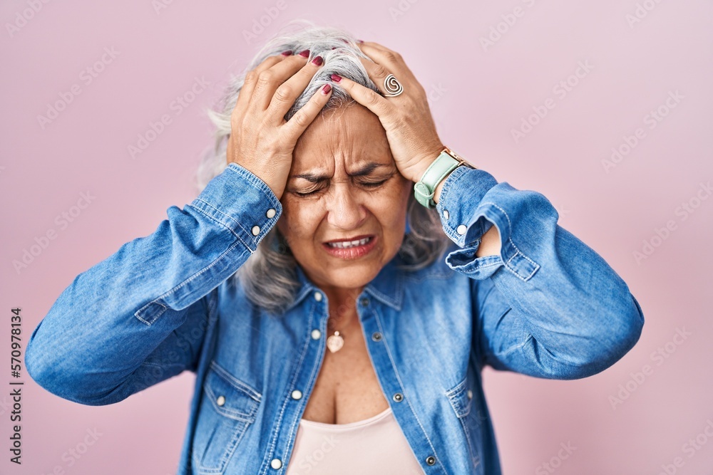 Poster middle age woman with grey hair standing over pink background suffering from headache desperate and 