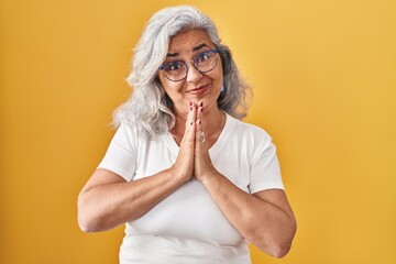 Middle age woman with grey hair standing over yellow background praying with hands together asking for forgiveness smiling confident.