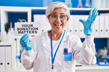 Middle age woman with grey hair working at scientist laboratory holding your donation matters banner celebrating victory with happy smile and winner expression with raised hands