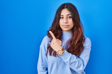 Hispanic young woman standing over blue background pointing with hand finger to the side showing advertisement, serious and calm face