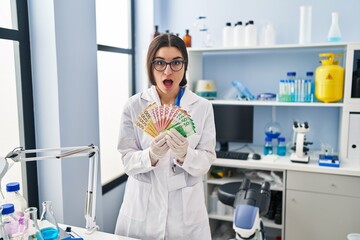Young hispanic woman working at scientist laboratory holding money banknotes afraid and shocked with surprise and amazed expression, fear and excited face.