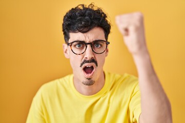 Hispanic man wearing glasses standing over yellow background angry and mad raising fist frustrated and furious while shouting with anger. rage and aggressive concept.